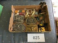 Overview of box containing four metal belt buckles with engraved designs, several vintage toy figurines in varied colors, and a bag with dice and poker chips.