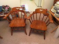 Two maple captain's chairs with curved backs and spindle supports placed side by side on carpeted floor.