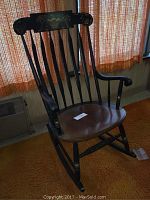 Front angled photo showing black spindle back rocking chair with decorative top rail and varnished wood seat, placed on orange carpet with curtain background.