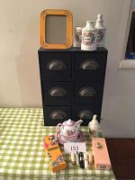 Full view of the black 6-drawer storage cabinet on a green and white checkered tablecloth with tea set, perfume bottles, boxes, and yellow photo frame on and around it.