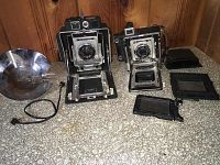 Two vintage cameras on a countertop: Busch Pressman Model D (left) and Graflex Crown Graphic (right) with parts and accessories.