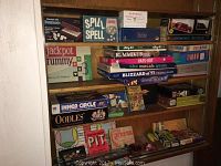 Wide view of a wooden shelving unit filled with various board games including Double Six Dominoes, Spill and Spell, Jackpot Rummy, Rummikub, and several others, mostly vintage boxes.