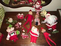 Top down view of assorted Santa Claus and elf figurines on wooden table, showing a mix of flocked fabric, celluloid heads, and painted features.