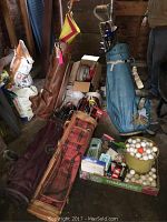 Photo showing three golf bags (brown leather, red plaid, blue) with assorted golf clubs and boxes on the floor around them.
