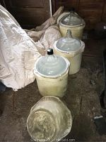 Three glass carboys each seated within ceramic fermentation vessels placed on a dusty floor. One ceramic vessel lid is removed and placed in front showing the interior. The ceramic vessels are cream colored with dirt and use marks.