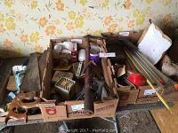 Cardboard boxes holding various hand tools including saws with wooden handles, a rusted metal tool, and small hardware parts.