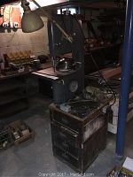 Wide angle photo showing the band saw mounted on a wooden cabinet base in a basement workshop environment, with drawers and some boxes visible nearby.