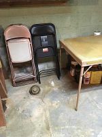 Six metal folding chairs, three beige and three black, leaning against wall next to folding card table.