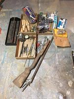 View of full lot showing wooden furniture legs, plumbing parts in plastic packaging, metal hardware, vintage gun stocks, and miscellaneous tools spread on floor with a metal toolbox or tray