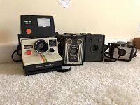 Five vintage cameras including a Polaroid OneStep in front, two Kodak Brownie Six-20 cameras, a Kodak Brownie Holiday, and a Polaroid Land Camera 800 in case at back.