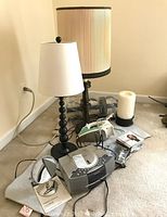 Photo showing two lamps, one black spindle-style with white shade and another tall wooden-style with beige pleated shade, along with a pillar candle and power strip on carpeted floor against wall.