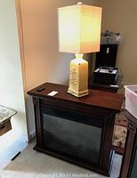 Dark wood electric fireplace heater with black front panel and beige ceramic lamp on top, both sitting on carpeted floor in room corner.