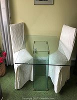Square glass table with cubic base and two white covered chairs, positioned near window, green carpet background.