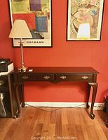Full view of wooden sofa table with traditional legs, two drawers, dark finish, placed against a red wall with framed artwork above. Faux brass table lamp sits on the left side of the table.