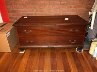 Front view of closed vintage wooden hope chest showing polished wood finish, decorative edge trim, and brass-tone handles on a hardwood floor beside a brick wall.