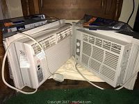 Photo showing two white window air conditioners on a surface, cords visible, with original tags attached.