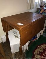 Wooden drop leaf table with turned legs, showing top surface and side leaf in folded-down position, with visible wear and scratches.