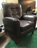 Brown leatherette recliner chair with button tufted backrest, rounded armrests, and wooden legs in a room with a TV and green rug.