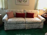 Front view of floral patterned sofa with three cushions in a room with green carpet and floral wall art.
