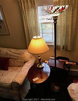 Side view of the oval wood veneer side table with faux brass table lamp and Tiffany style stained glass floor lamp behind, placed next to a floral upholstered couch and window with drapes.