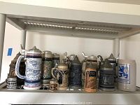 Multiple beer steins arranged on a plastic shelf, including various sizes, materials, and decorative reliefs.
