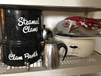 14 inch black speckled clam pot with upper section labeled 'Steamed Clams' and lower section labeled 'Clam Broth', along with two metal perk pots, CorningWare crockpot, and folded outdoor textiles on a shelf.