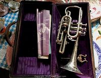 Open trumpet case showing the silver-plated Bruno NYC trumpet with detailed floral engravings resting inside the purple fabric-lined case along with some folded sheet music.