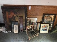 Overview of lot showing various framed artwork, mirror, wooden thermometer, folk art chair, and horseshoes on the floor.