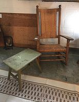 Primitive oak wood rocking chair with woven rattan seat and backrest along with a small painted side stool nearby.