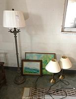 Photo showing 72-inch tall floor lamp with decorative base and pleated shade, two small brass table lamps with cream shades, and two framed watercolor paintings leaning against wall