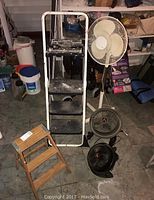 Photo showing three fans and two step ladders in used condition, placed on a garage floor.