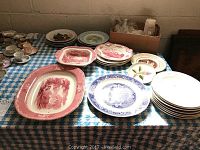 Wide view of various reproduction and commemorative china plates laid out on a checkered table cloth along with assorted clear cut glass cups on the far right side and a box behind the plates containing additional items.