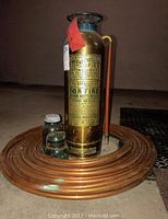 Full view of brass fire extinguisher standing in the center of coiled copper pipe with two small blue glass jars in front.