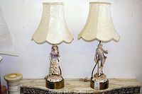Two Victorian porcelain couple lamps side by side on a marble surface, each with scalloped cream fabric lampshades and detailed hand-painted porcelain bases depicting a man and woman.