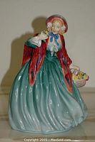 Front three-quarter view of glazed bone china figurine in green skirt and red cloak holding basket of flowers.
