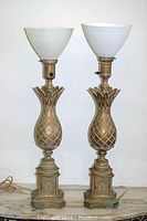 Pair of metal pineapple motif table lamps with milk glass shades on a marble surface against a white background