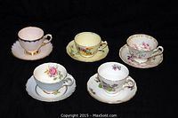 Five assorted fine china cups and saucers on black background, all featuring floral designs and gold trim.