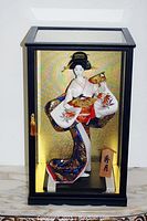 Front view of Japanese Geisha doll in glass showcase with black frame and gold background