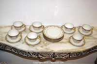 Full view of 7 cups with gold trimmed handles and saucers, 5 lunch plates stacked, all placed on white marble surface with decorative black and gold trim below.