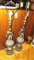 Photo showing both vintage bronze tone table lamps with cherub figures on a globular ornate base, unlit, set against a wood paneled wall