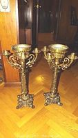 Photo showing two large bronze tone metal candelabras with ornate cast metal design, four arms each, and central candle holders.