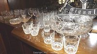 Wide view showing multiple crystal glasses including highballs, rock glasses, ashtrays, and a large footed bowl on a dark surface.