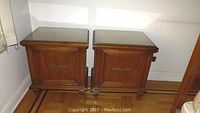Pair of fruitwood end tables with glass tops and bronze-tone hardware, closed doors showing the front panel and handles.