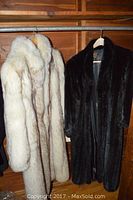 Both fur coats hanging on rod in closet; one white/silver fox, one dark mink