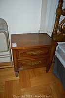 Closed end table showing the front with two drawers and bronze-tone hardware, medium brown finish, on wooden floor next to bed and folding chair