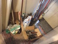 Long-handled garden tools including a shovel, post hole digger, bagged soil or fertilizer, and smaller hand tools in a cardboard box.