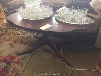 Full view of oval wooden coffee table with curved pedestal base and tripod legs, with decorative glassware on top, showing overall shape and structure.