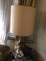 Two vintage brass table lamps with beige fabric shades on wooden side table near window with drapes.
