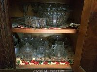 Glass bowls, candy dishes, trays, plates displayed on two shelves inside a wooden cabinet, showing a variety of clear glassware.