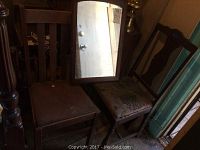 Two wooden side chairs with visibly worn or damaged seat coverings next to a rectangular mirror with wooden frame in a dimly lit room.